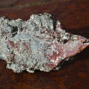 May include: A rough, irregularly shaped rock with a mix of gray and reddish-brown hues. The surface has a textured, weathered appearance, suggesting a natural mineral formation. The rock sits on a dark brown surface.