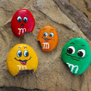 May include: Four painted rocks in red, orange, yellow, and green, each with a smiling face and a white "m" design. The rocks are on a textured, brown stone surface. The rocks are painted to resemble M&M's sweets.