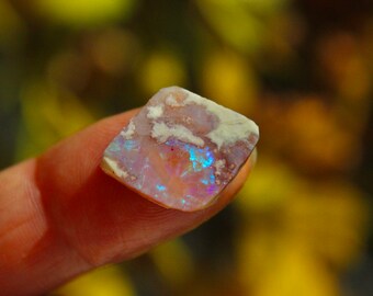 May include: A small, rough-cut piece of opal with a white, pink, and blue iridescent sheen. The opal is held in a hand.
