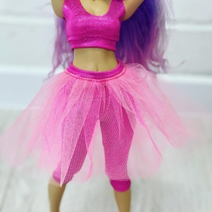 May include: A doll wearing a pink outfit. The outfit includes a sparkly crop top, a tutu-style skirt, and capri leggings. The doll has purple hair.