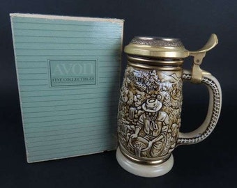 May include: A vintage Avon Fine Collectibles beer stein with a detailed relief scene of a prospector. The stein has a brass-colored lid and handle, and a cream-colored base. The box is light green with the Avon logo.