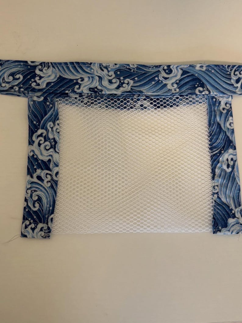 May include: A rectangular laundry bag with a white mesh body and a blue fabric border. The fabric border features a repeating pattern of white waves on a dark blue background. The bag is designed for washing delicate items.