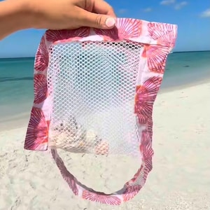 May include: A mesh beach bag with a pink seashell pattern. The bag is filled with seashells and held up against a backdrop of a sandy beach and blue ocean.