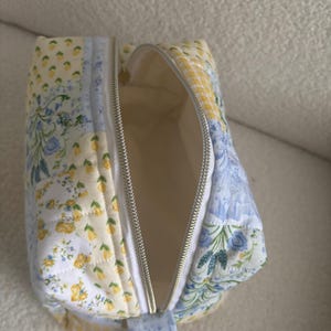 K&ouml;nnte beinhalten: Ge&ouml;ffnete, gesteppte Kosmetiktasche mit floralem Patchwork-Design in Gelb und Blau. Die Tasche hat einen silbernen Rei&szlig;verschluss und einen passenden Stoff-Zuglasche. Die Innenseite ist hellcremefarben.