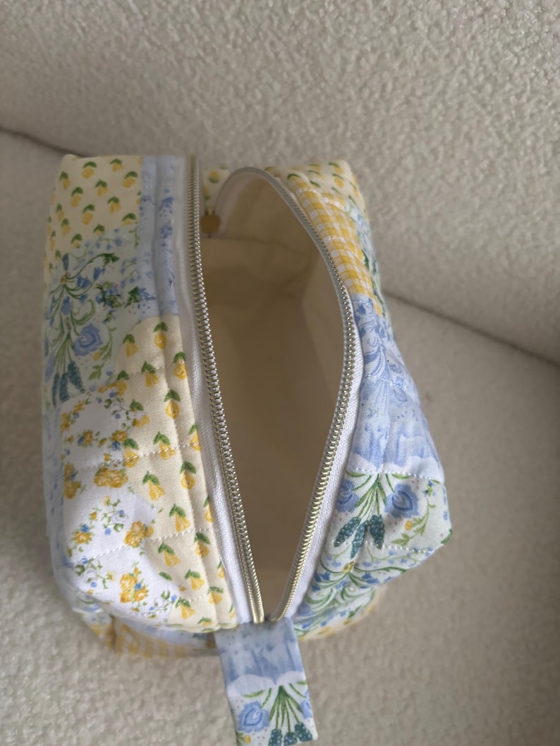 K&ouml;nnte beinhalten: Ge&ouml;ffnete, gesteppte Kosmetiktasche mit floralem Patchwork-Design in Gelb und Blau. Die Tasche hat einen silbernen Rei&szlig;verschluss und einen passenden Stoff-Zuglasche. Die Innenseite ist hellcremefarben.