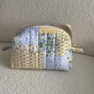May include: A quilted cosmetic bag with a patchwork design. The bag features a mix of floral, gingham, and solid fabric panels in yellow, blue, and white. It has a silver zipper and small fabric loops on the sides.