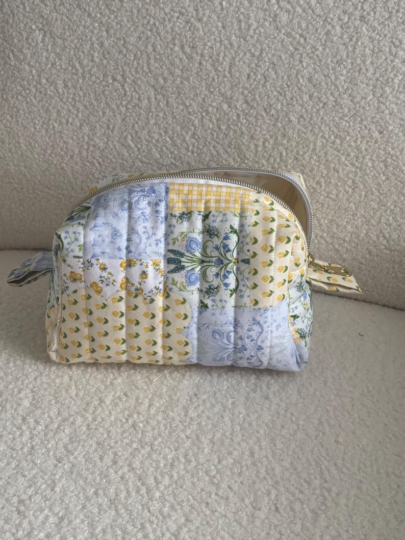 K&ouml;nnte beinhalten: Eine gesteppte Kosmetiktasche mit Patchwork-Design. Die Tasche hat eine Mischung aus floralen, Vichy- und einfarbigen Stoffbahnen in Gelb, Blau und Wei&szlig;. Sie hat einen silbernen Rei&szlig;verschluss und kleine Stoffschlaufen an den Seiten.