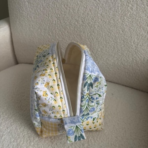 K&ouml;nnte beinhalten: Eine Kosmetiktasche aus Patchwork mit einem wei&szlig;en Rei&szlig;verschluss. Die Tasche besteht aus verschiedenen Stoffen mit gelben, blauen und wei&szlig;en Blumenmustern sowie einem gelben Vichymuster. Der Innenraum ist cremefarben.