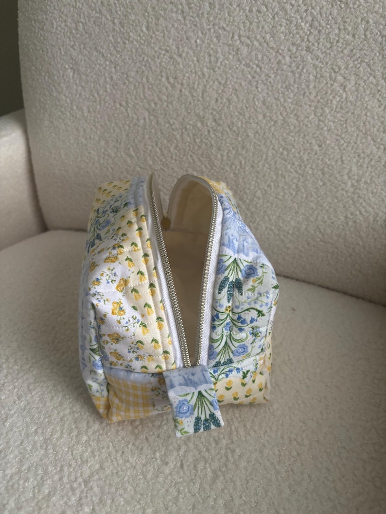 K&ouml;nnte beinhalten: Eine Kosmetiktasche aus Patchwork mit einem wei&szlig;en Rei&szlig;verschluss. Die Tasche besteht aus verschiedenen Stoffen mit gelben, blauen und wei&szlig;en Blumenmustern sowie einem gelben Vichymuster. Der Innenraum ist cremefarben.
