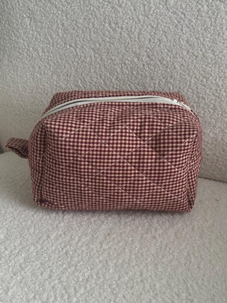 K&ouml;nnte beinhalten: Eine gesteppte Kosmetiktasche mit burgunderrotem und rosa Vichy-Muster und einem wei&szlig;en Rei&szlig;verschluss. Die Tasche hat eine kleine Stoffschlaufe. Ma&szlig;e in Zentimetern.