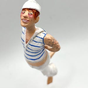 Könnte beinhalten: Eine handbemalte Figur eines Menschen, der einen weißen Matrosenhut, ein blau-weiß gestreiftes Hemd und eine weiße Hose trägt. Die Figur hat rote Haare und ein Tattoo auf dem Arm.