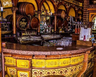 May include: An interior shot of a classic bar with wooden barrels, bottles, and a curved bar. The bar is decorated with ornate yellow and brown tiles and a wooden stool. The text "HIGH CLASS HISKIES. DIRECT IMPORTERS" is visible.
