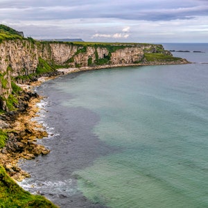 May include: A view of a rocky coastline with green grass and cliffs. The ocean is a light blue-green color and there are small islands in the distance.