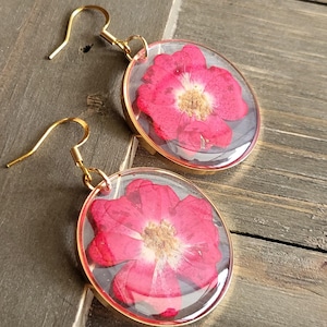 May include: A pair of gold earrings with a red pressed flower encased in clear resin. The earrings have a simple hook closure.