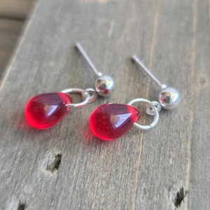 May include: A pair of earrings with teardrop-shaped, translucent red beads hanging from silver hoops and posts. The earrings have a polished surface and a simple design. The posts are silver with a small, round ball at the end. The earrings are a striking red colour.