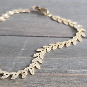 May include: A gold-colored necklace featuring a delicate leaf design. The necklace is made of small, detailed leaves linked together, creating a flowing, elegant look. The clasp is visible at the top, and the necklace rests on a wooden surface.