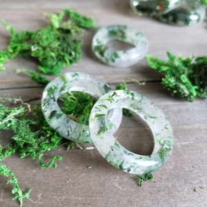 May include: Four clear resin rings with embedded green foliage. The rings are stacked on a wooden surface with green moss.