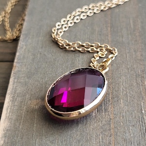 May include: A gold-toned necklace with a large, faceted, oval-shaped, deep magenta pendant. The pendant is set in a gold bezel and hangs from a delicate gold chain. The necklace rests on a weathered, gray wooden surface.