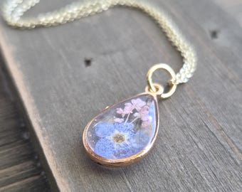 Puede incluir: Un collar dorado con un colgante en forma de lágrima. El colgante contiene flores secas azules y rosas encapsuladas en resina transparente. La cadena tiene un diseño delicado y tejido. El collar descansa sobre una superficie de madera gris envejecida.
