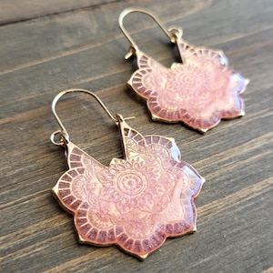 Puede incluir: Un par de pendientes de aro dorados con un diseño de mandala rosa y dorado. Los pendientes están hechos de metal y tienen un diseño delicado e intrincado.