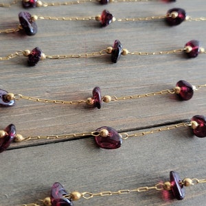 May include: A gold chain necklace with multiple strands of small, dark red gemstone beads. The necklace is laid out on a wooden surface.