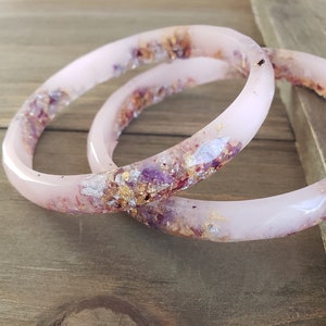 Puede incluir: Dos pulseras de resina rosa con escamas doradas y moradas incrustadas. Las pulseras están apiladas una encima de la otra.