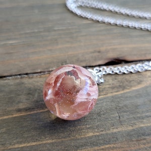 May include: A pink and white resin sphere pendant with a silver chain. The sphere has a swirling pattern and is translucent.