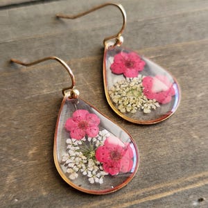 May include: Teardrop earrings with gold-coloured hooks. Each earring has a clear resin enclosure with pink flowers and white filler flowers. The earrings are set against a wooden background.