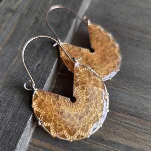 May include: A pair of handcrafted earrings with a unique design. The earrings feature a half-moon shape with a detailed, golden-brown pattern encased in a clear resin. The ear wires are silver-colored, adding a contrasting metallic accent. The earrings are displayed on a wooden surface.