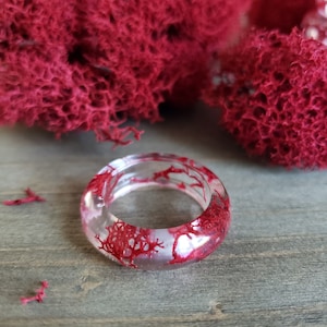 May include: A clear resin ring with red dried botanicals embedded inside. The ring is on a wooden surface.