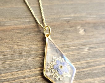 Puede incluir: Un collar de cadena dorada con un colgante de diamante transparente que contiene una flor morada prensada y flores blancas.