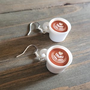 May include: A pair of white coffee cup earrings with brown coffee and white latte art. The earrings have silver hooks.