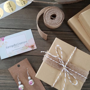 Puede incluir: Una peque&ntilde;a caja marr&oacute;n envuelta con cuerda y una cinta dorada. La caja est&aacute; sobre una superficie de madera con una tarjeta de visita para Earrings ByL Creations y un par de pendientes florales en una tarjeta marr&oacute;n.