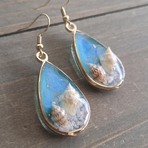 May include: Teardrop-shaped earrings with a gold-colored frame. Each earring features a light blue background with small seashells and sand-like elements encased in clear resin. The earrings have gold-colored fish hook ear wires.