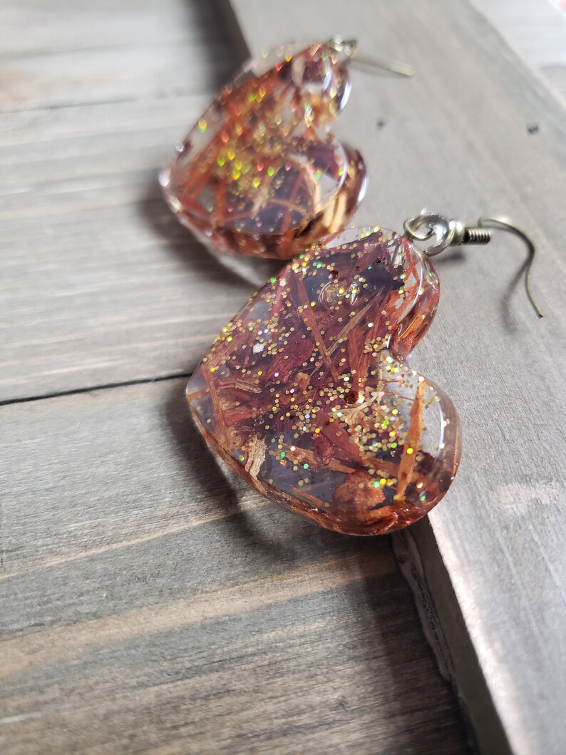 May include: A pair of heart-shaped resin earrings with gold glitter and brown and red dried flowers embedded inside. The earrings have silver hooks.