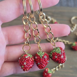 以下が含まれることがあります： ゴールドトーンのフープピアス、イチゴのチャーム付き。 イチゴは赤と緑のラインストーンで装飾され、フープはゴールドチェーンでチャームに接続されています。 楽しくて気まぐれなアクセサリーです。