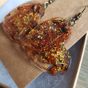 May include: A pair of heart-shaped resin earrings with gold glitter and dried flowers embedded inside. The earrings are attached to gold-toned hooks.