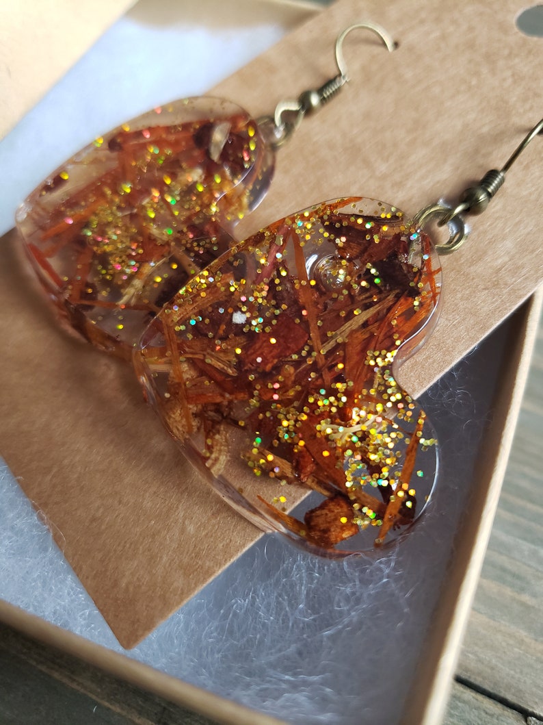 May include: A pair of heart-shaped resin earrings with gold glitter and dried flowers embedded inside. The earrings are attached to gold-toned hooks.