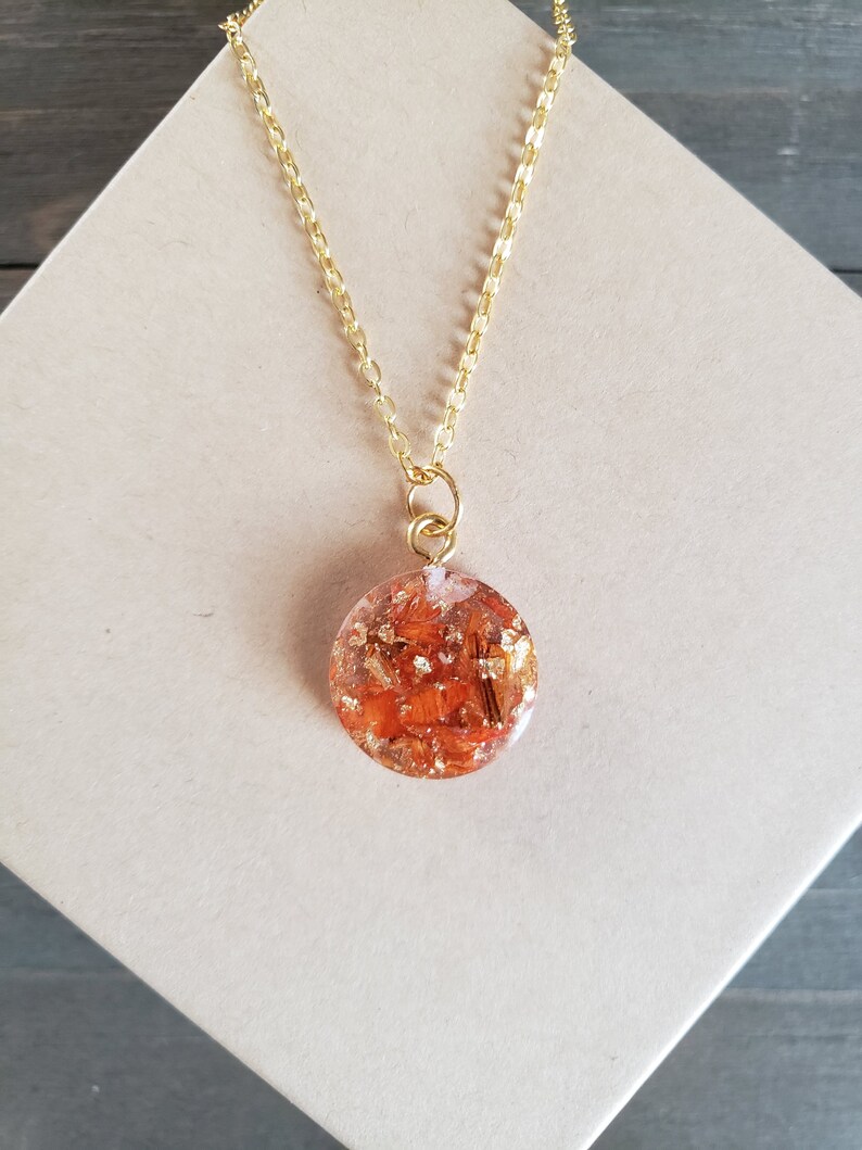 May include: A gold chain necklace with a round pendant featuring orange and gold flakes encased in clear resin.