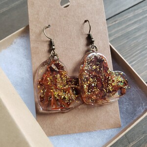 May include: A pair of heart-shaped resin earrings with gold glitter and dried flowers. The earrings are on a brown card with a hole for hanging.