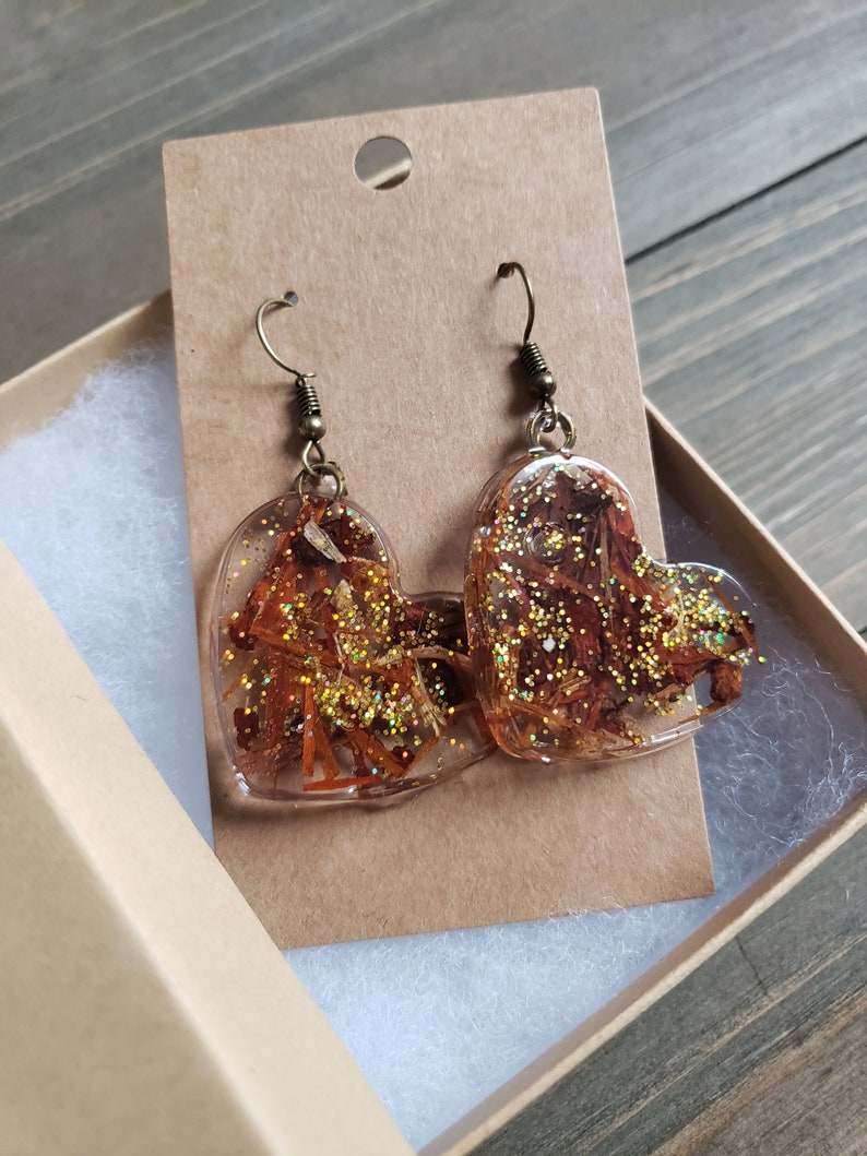 May include: A pair of heart-shaped resin earrings with gold glitter and dried flowers. The earrings are on a brown card with a hole for hanging.