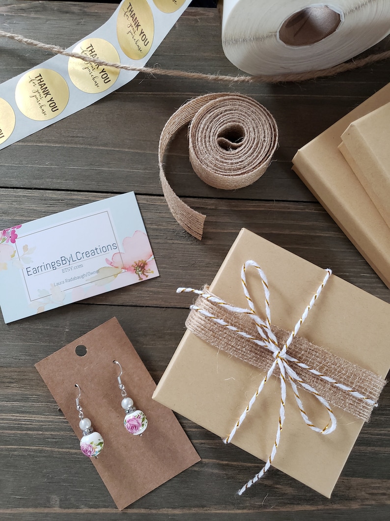 May include: A small brown box wrapped with twine and a gold ribbon. The box is sitting on a wooden surface with a business card for Earrings ByL Creations and a pair of floral earrings on a brown card.