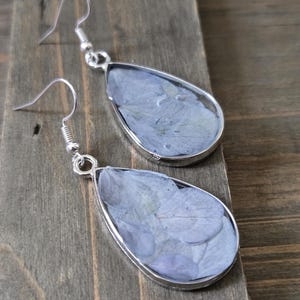 May include: A pair of teardrop earrings with silver frames. Each earring features a light blue floral design set in clear resin. The earrings hang from silver fish hook wires. The earrings are displayed on a wooden surface.