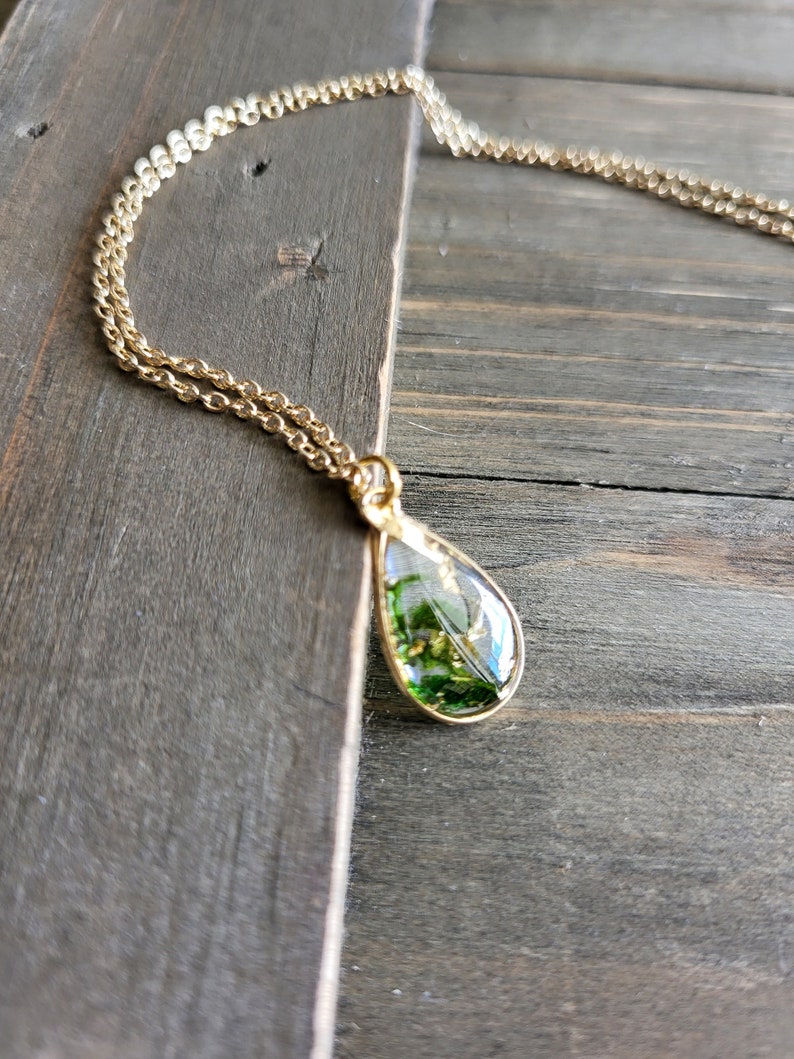May include: A gold chain necklace with a teardrop-shaped pendant. The pendant is filled with green and gold pressed flowers and leaves.
