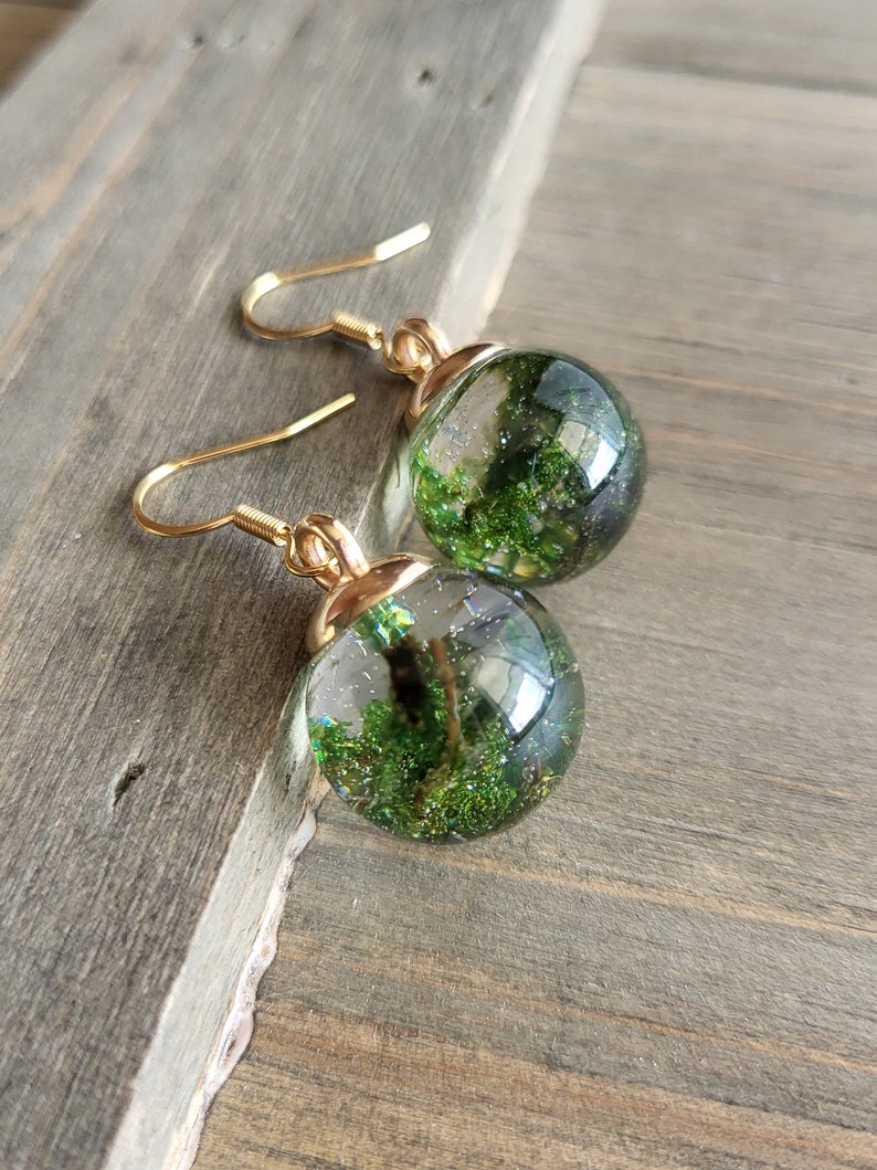 May include: Gold earrings with a green glitter ball charm. The ball is filled with green glitter and has a clear resin finish.