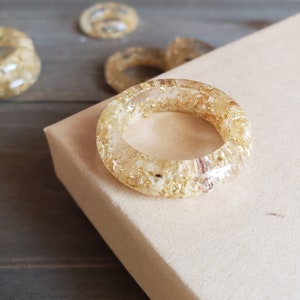 Op de afbeelding: Een gouden harsring met ingebedde gouden vlokken. De ring ligt op een lichtbruin oppervlak.
