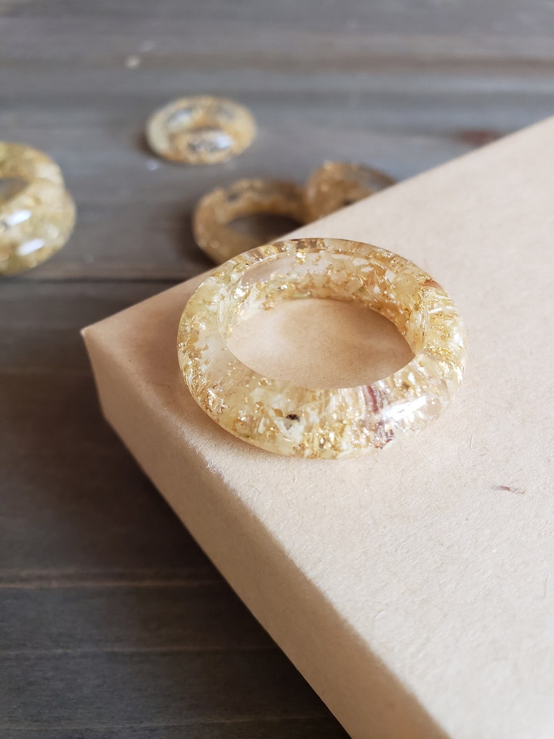 Op de afbeelding: Een gouden harsring met ingebedde gouden vlokken. De ring ligt op een lichtbruin oppervlak.