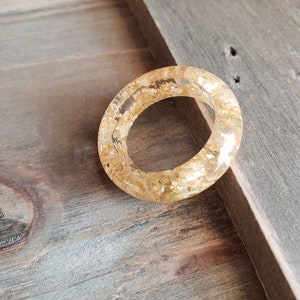 Op de afbeelding: Een heldere ring met gouden vlokken ingebed in de hars. De ring ligt op een houten oppervlak.
