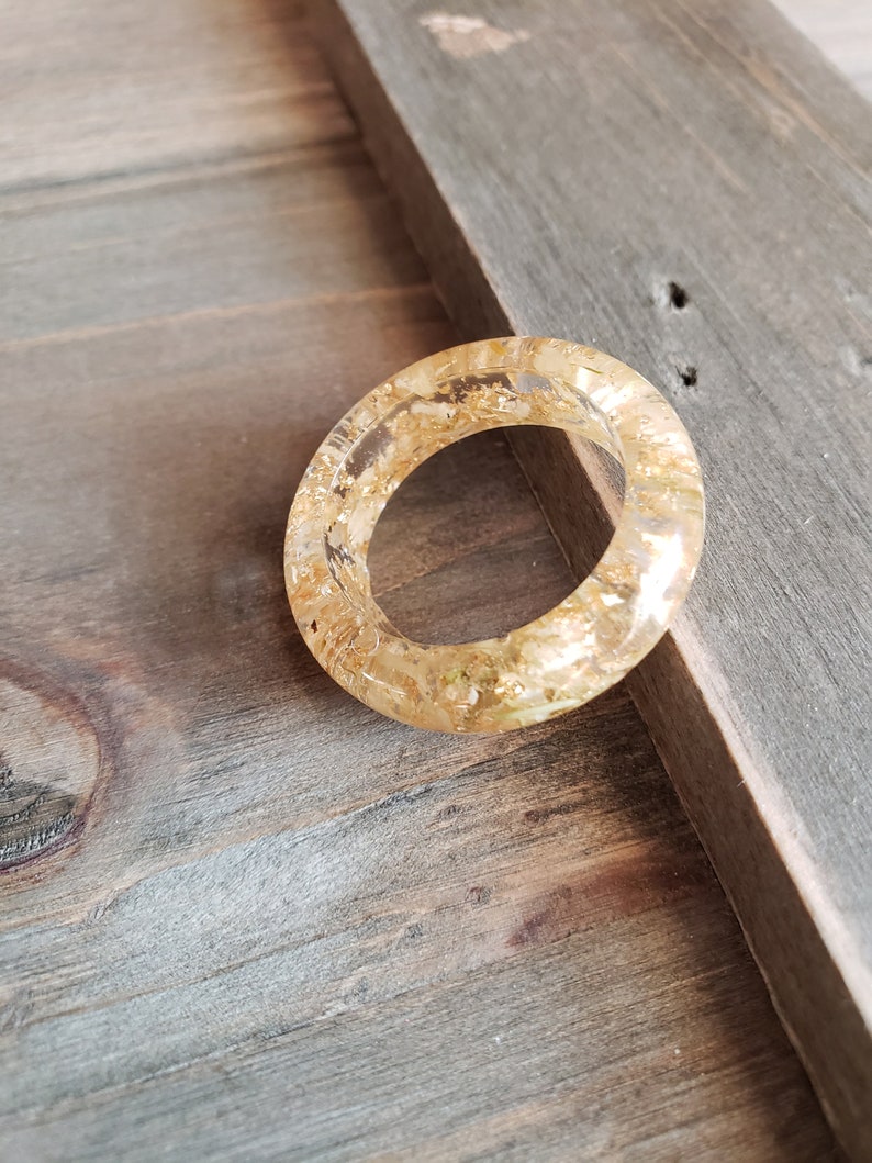 Op de afbeelding: Een heldere ring met gouden vlokken ingebed in de hars. De ring ligt op een houten oppervlak.