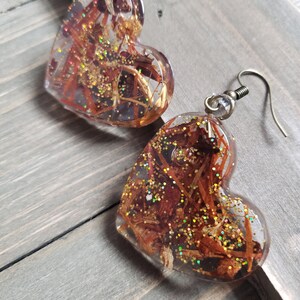 May include: A pair of heart-shaped resin earrings with gold glitter and brown wood-like inclusions. The earrings have silver hooks.